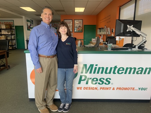 Minuteman Press Franchise Owners Ramon and Alana stand in front of a desk with the Minuteman logo