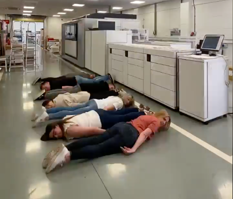 Tadazz employees laying on the pressroom floor in front of a printer press